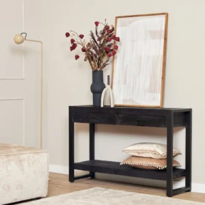 De Sidetable Boaz Black 120 cm staat tegen een beige muur, met daarop twee vazen - één met gedroogde bloemen - en ingelijste abstracte kunst. Twee kussens rusten op de onderste plank, met een staande lamp en een beige voetenbank in de buurt.