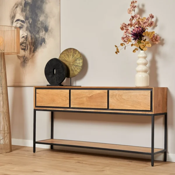 De Sidetable RichWood | 150 cm, een moderne houten console met drie laden en zwarte metalen poten, staat tegen een lichte muur met decor, een hoge vaas met bloemen en ronde sculpturen. Vlakbij hangen een schilderij en een geweven vloerlamp.