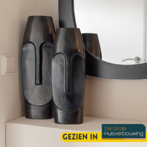 De Vaas Liv - Zwart | Large met een modern abstract gezichtsontwerp staat op een hoekplank naast een ronde spiegel. In de rechterbenedenhoek van de afbeelding staat de tekst "GEZIEN IN De Grote Huisverbouwing".