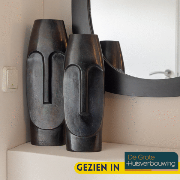 De Vaas Liv - Zwart | Large met een modern abstract gezichtsontwerp staat op een hoekplank naast een ronde spiegel. In de rechterbenedenhoek van de afbeelding staat de tekst "GEZIEN IN De Grote Huisverbouwing".