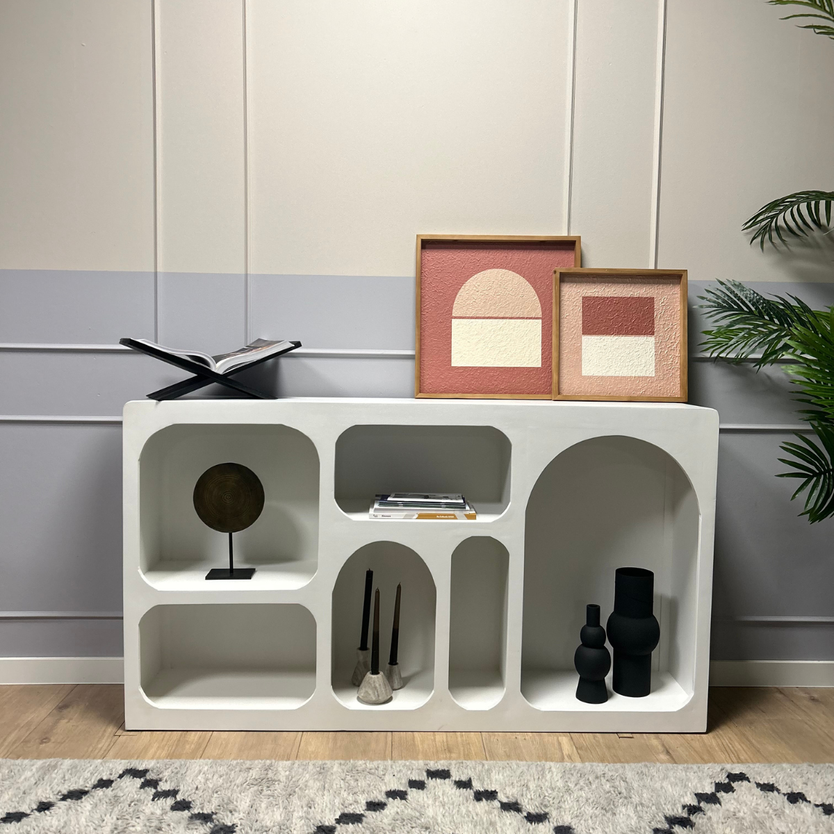 De Dressoir Caya Wit, versierd met abstracte kunst in houten frames, zwarte vazen, kwasten, een rond beeldhouwwerk en een open boek, staat tegen een licht gepaneelde muur naast een groene plant.