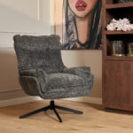 De Fauteuil Vera Dark Grey, een pluche draaifauteuil met gestructureerde stof, staat op een houten vloer naast een houten plank met daarop een vaas met roze bloemen en een schilderij van een vrouwengezicht op de achtergrond.