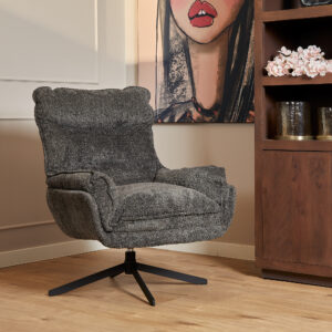 De Fauteuil Vera Dark Grey, een pluche draaifauteuil met gestructureerde stof, staat op een houten vloer naast een houten plank met daarop een vaas met roze bloemen en een schilderij van een vrouwengezicht op de achtergrond.