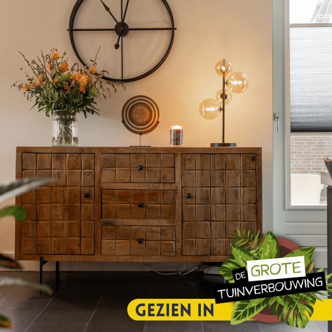 De Dressoir Brandy | 160 cm, met een geruit patroon, staat tegen de muur met een vaas, bord en lamp erop. Een grote klok hangt erboven. Overlay tekst: "Gezien in De Grote Tuinverbouwing.