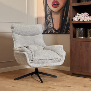 De Fauteuil Vera Lichtgrijze draaifauteuil met pluche kussens staat op een houten vloer naast een boekenplank en een ingelijst kunstwerk van een vrouwengezicht. Roze bloemen en glasdecor maken de gezellige, moderne omgeving compleet.
