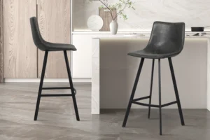 Twee Barstoel 'Ozan' PU Zwart krukken (set van 2) met slanke metalen poten staan in een keuken met lichte kasten, marmeren werkbladen en een minimalistische inrichting met een vaas van groene takken.