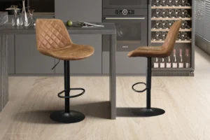 Twee Barstoel 'Madrid' PU Ote Topper 02 Cognac barkrukken (2 per doos) met gewatteerde bruine zittingen en zwart metalen onderstel staan bij een grijs keukeneiland, in een moderne keuken met grijze kasten, een wijnkoelkast en een oven.