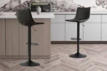 Twee 'Trevi' zwarte PU barkrukken (set van 2) met voetsteunen staan bij een licht houten keukeneiland. Witte kastjes, een marmeren achterwand en een grijze vaas sieren de achtergrond, terwijl de vloer een chevron houtpatroon heeft.