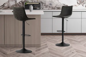 Twee 'Trevi' zwarte PU barkrukken (set van 2) met voetsteunen staan bij een licht houten keukeneiland. Witte kastjes, een marmeren achterwand en een grijze vaas sieren de achtergrond, terwijl de vloer een chevron houtpatroon heeft.