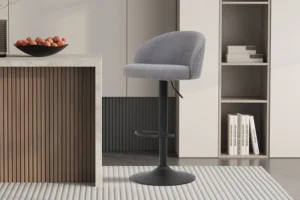 Een moderne keuken toont de Barstoel 'Arda' Bouclé Lichtgrijs (2 per doos) met een zwart metalen onderstel naast een marmeren aanrecht. Planken met boeken en decor verschijnen op de achtergrond, terwijl een schaal met fruit het aanrecht siert.