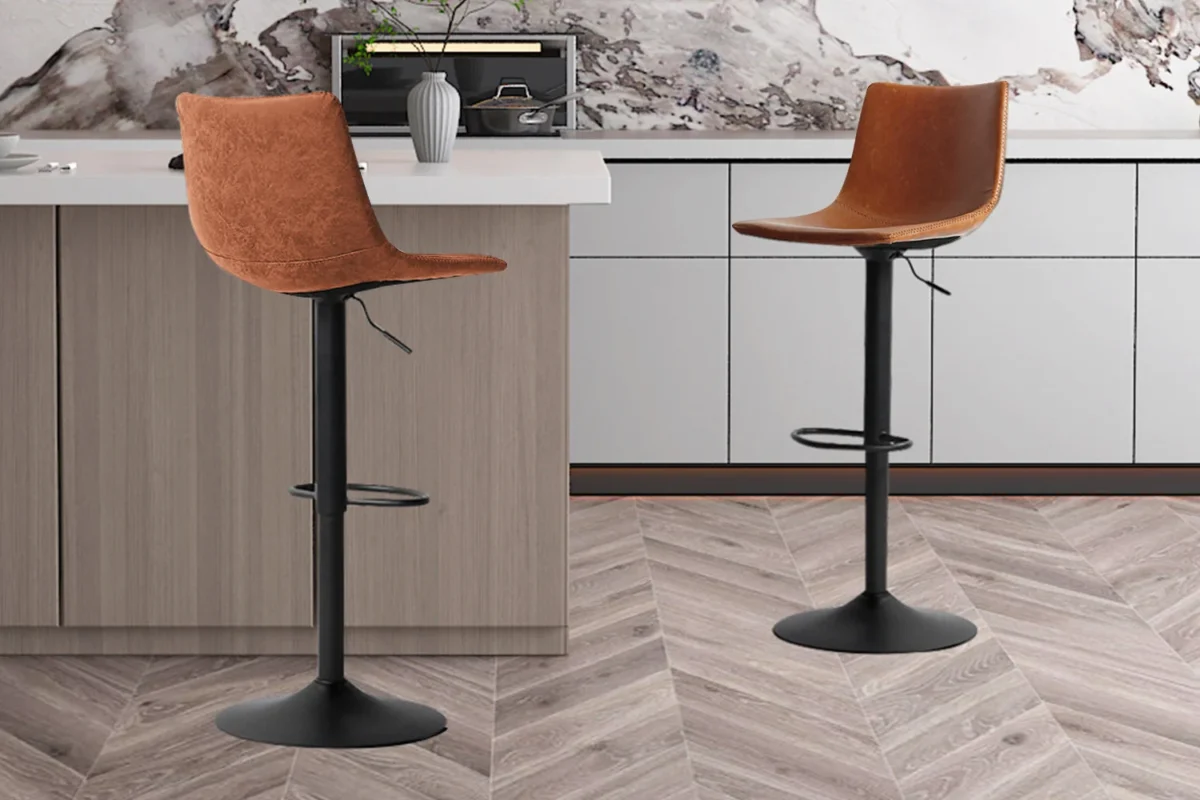 Twee 'Trevi' PU Cognac barkrukken (set van 2) met zwart metalen onderstel staan naast een lichthouten keukeneiland met witte werkbladen en kasten. De vloer heeft een visgraatmotief.