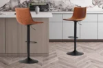 Twee 'Trevi' PU Cognac barkrukken (set van 2) met zwart metalen onderstel staan naast een lichthouten keukeneiland met witte werkbladen en kasten. De vloer heeft een visgraatmotief.