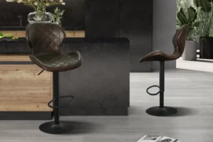 Twee Barstoel 'Malaga' PU Ote Topper 05 Donkerbruin krukken (set van 2) met zwart metalen onderstel staan in een stijlvolle keuken met houten kasten, een zwart aanrechtblad en planten. Een open tijdschrift rust op de lichtgrijze tegelvloer.
