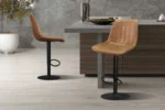 Twee 'Barcelona' PU Ote Topper 02 Cognac barkrukken (set van 2) met zwart metalen onderstel staan bij een modern keukeneiland met een grijs stenen aanrechtblad, lichthouten vloeren, grijze stenen muren en een plant in pot.