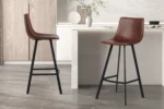 Twee Barstoel 'Ozan' PU Bruin barkrukken (set van 2) met zwart metalen poten staan in een strakke keuken met lichte kasten, marmeren aanrechtblad en een decoratieve vaas met takken op het aanrecht.