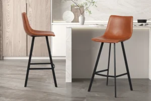 Twee Barstoel 'Ozan' PU Cognac krukken (verkocht als set van 2) met zwart metalen poten staan in een moderne keuken met licht hout en marmeren afwerking. Op de achtergrond staan decoratieve vazen en een plant op het aanrecht.