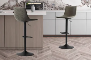 Twee Barstoel 'Trevi' PU Grijs barkrukken (set van 2) met draaibare zittingen en zwart onderstel staan bij een lichthouten keukeneiland met wit aanrechtblad, aangevuld met kasten en een decoratieve vaas op de achtergrond.