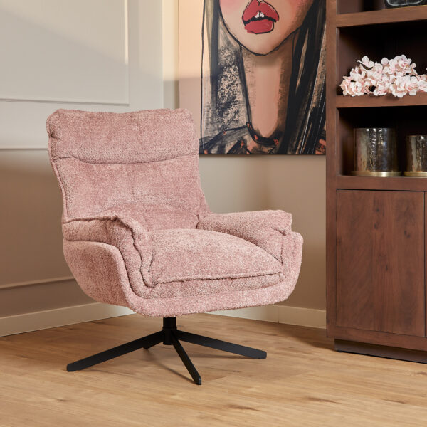 De Fauteuil Vera Roze draaifauteuil staat op een houten vloer naast een versierde boekenplank, met een groot kunstwerk van een vrouwengezicht gedeeltelijk zichtbaar op de muur erachter.