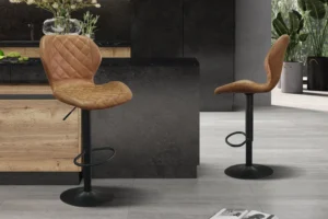 Twee Barstoel 'Malaga' PU Ote Topper 02 Cognac barkrukken, met bruin gewatteerde zittingen en zwart metalen onderstellen, staan in een stijlvolle keuken met houten en donkere kasten. Naast hen ligt een krant op de grijze tegelvloer.