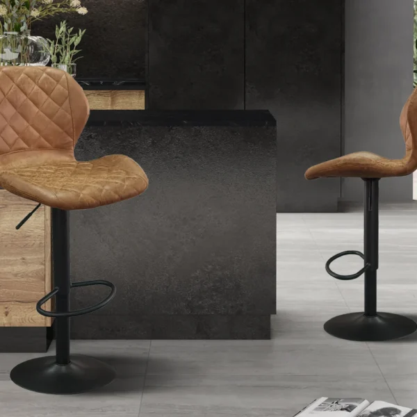 Twee Barstoel 'Malaga' PU Ote Topper 02 Cognac barkrukken, met bruin gewatteerde zittingen en zwart metalen onderstellen, staan in een stijlvolle keuken met houten en donkere kasten. Naast hen ligt een krant op de grijze tegelvloer.