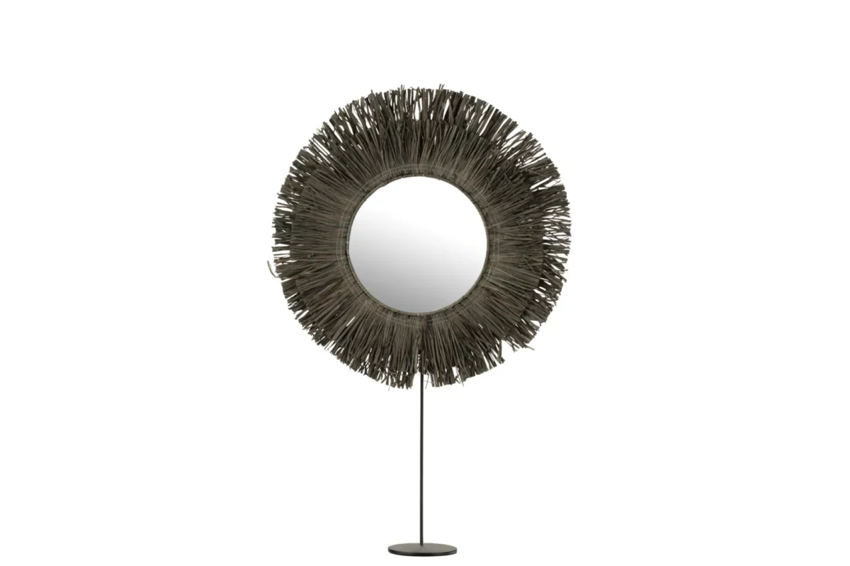 De Mirror On Foot Round Reed Raffia Grey heeft een ronde spiegel met een lijst in sunburst-stijl van donkere, slanke stokjes, gemonteerd op een slanke standaard met een ronde basis, tegen een witte achtergrond.