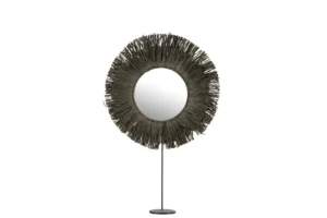 De Mirror On Foot Round Reed Raffia Grey heeft een ronde spiegel met een lijst in sunburst-stijl van donkere, slanke stokjes, gemonteerd op een slanke standaard met een ronde basis, tegen een witte achtergrond.