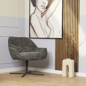 De Fauteuil Mila Donkergrijs staat naast een lichtbeige voetenbank met een vaas met takken, voor een groot vrouwenportret op een halflila, halfwitte muur met houten latten.