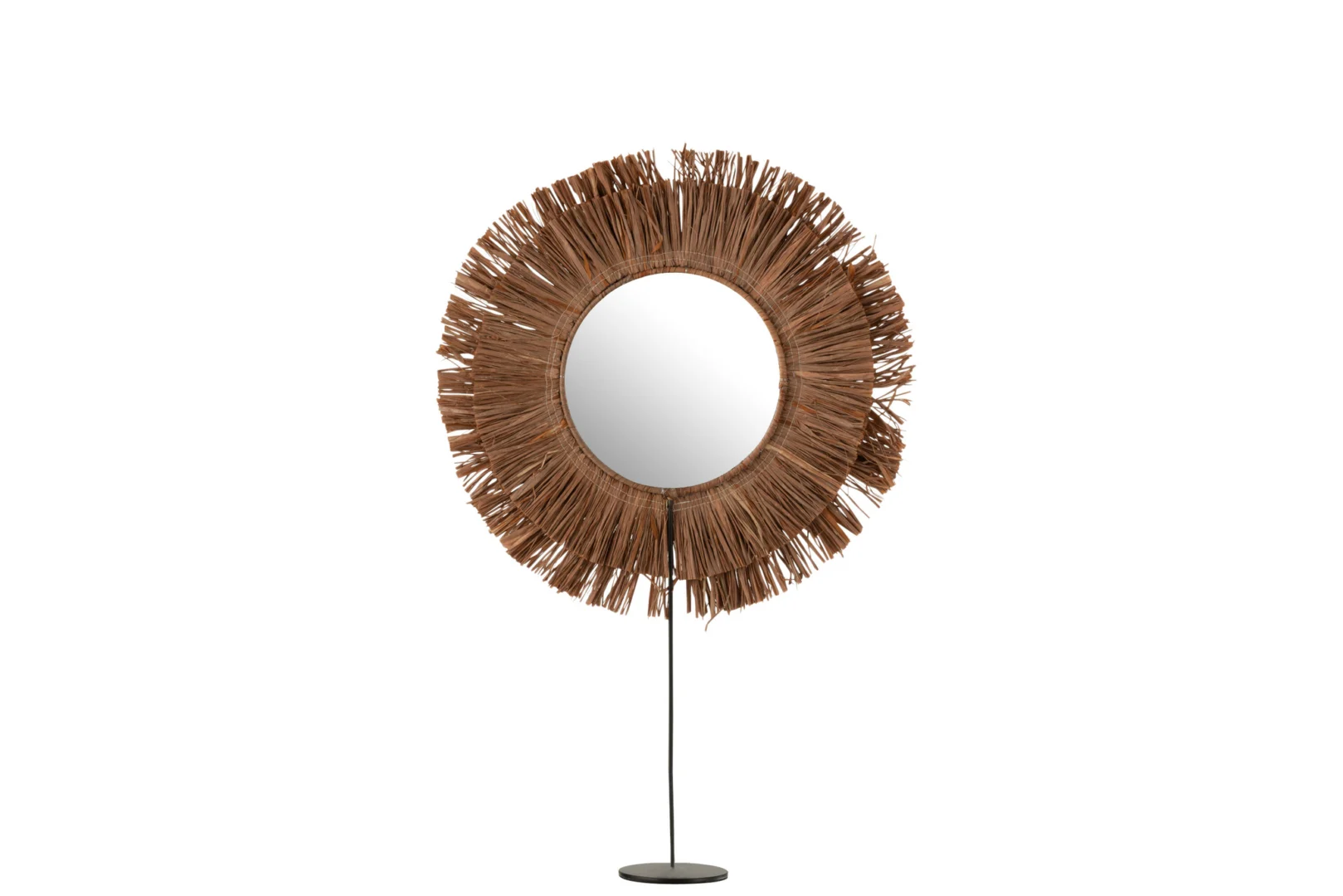 De Mirror On Foot Round Reed Raffia Brown heeft een ronde spiegel met een decoratieve sunburst lijst gemaakt van natuurlijk, stro-achtig raffia, gemonteerd op een dunne zwarte standaard met een ronde basis, afgebeeld tegen een witte achtergrond.