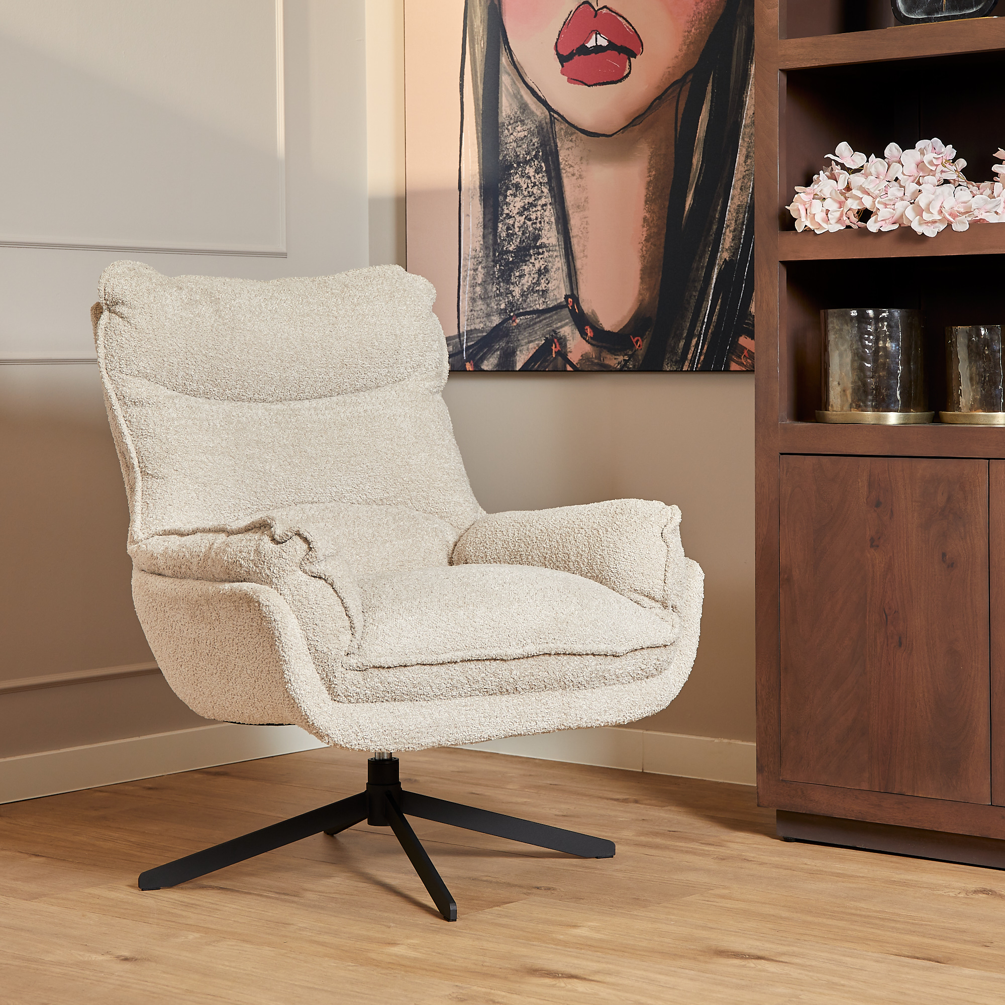 De Fauteuil Vera Off White, een beklede draaistoel in crème, staat op een houten vloer naast een bruine kast met decor en een schilderij van een vrouwengezicht aan de muur erachter.
