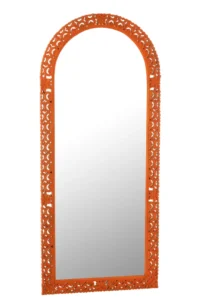De Mirror Carved Mumbai Mango Wood Mix is een hoge, gewelfde spiegel met een sierlijke oranje lijst met ingewikkelde bloemmotieven rond de randen, rechtopstaand tegen een witte achtergrond.