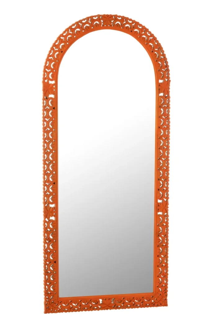 De Mirror Carved Mumbai Mango Wood Mix is een hoge, gewelfde spiegel met een sierlijke oranje lijst met ingewikkelde bloemmotieven rond de randen, rechtopstaand tegen een witte achtergrond.