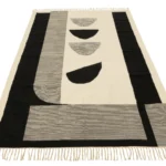 Het Vloerkleed Tokyo Poly Wit/Zwart is een rechthoekig vloerkleed met geometrische zwarte vormen en lijnen op een crèmekleurige achtergrond, met drie halve cirkels in het midden en franjes langs de kortere randen.