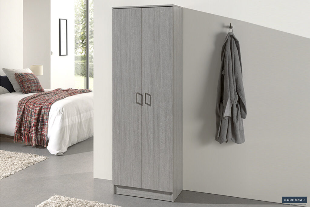 Een moderne slaapkamer met de Opbergkast 'Ray' met 2 deuren en 4 planken in grijs eiken, een badjas aan een wandhaak en een opgeruimd bed met een wit dekbed en geruite deken. Zonlicht stroomt door een raam op de achtergrond.