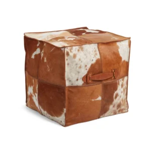De Kalfslederen Poef Vierkant Handvat V01 is een vierkante poef met bruin en wit patchwork van koeienhuid en een leren handvat aan één kant, afgebeeld tegen een effen witte achtergrond.
