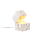 De TRIO Tafellamp TREASURE is een kubusvormige, witgekalkte houten lamp met een schuine open bovenkant die een gloeiende lamp binnenin onthult. De lamp staat op een plat oppervlak met een netsnoer dat uit de achterkant steekt.
