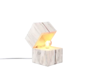 De TRIO Tafellamp TREASURE is een kubusvormige, witgekalkte houten lamp met een schuine open bovenkant die een gloeiende lamp binnenin onthult. De lamp staat op een plat oppervlak met een netsnoer dat uit de achterkant steekt.