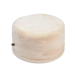 De Ronde Moccasin Poef 55x36cm V9 is een ronde, beige poef met een zachte, donzige textuur en een klein bruin stoffen label aan de zijkant, afgebeeld tegen een witte achtergrond.
