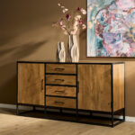De Dressoir Denver, gemaakt van mangohout en staal (165 cm), staat tegen een bruine muur. Twee hoge vazen met roze bloemen staan erop, met daarboven een kleurrijk abstract schilderij. De vloer is van licht hout.