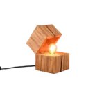 De TRIO Tafellamp TREASURE is een moderne houten lamp in de vorm van een open kubus, met een gloeilamp binnenin en een snoer dat uit de achterkant steekt.