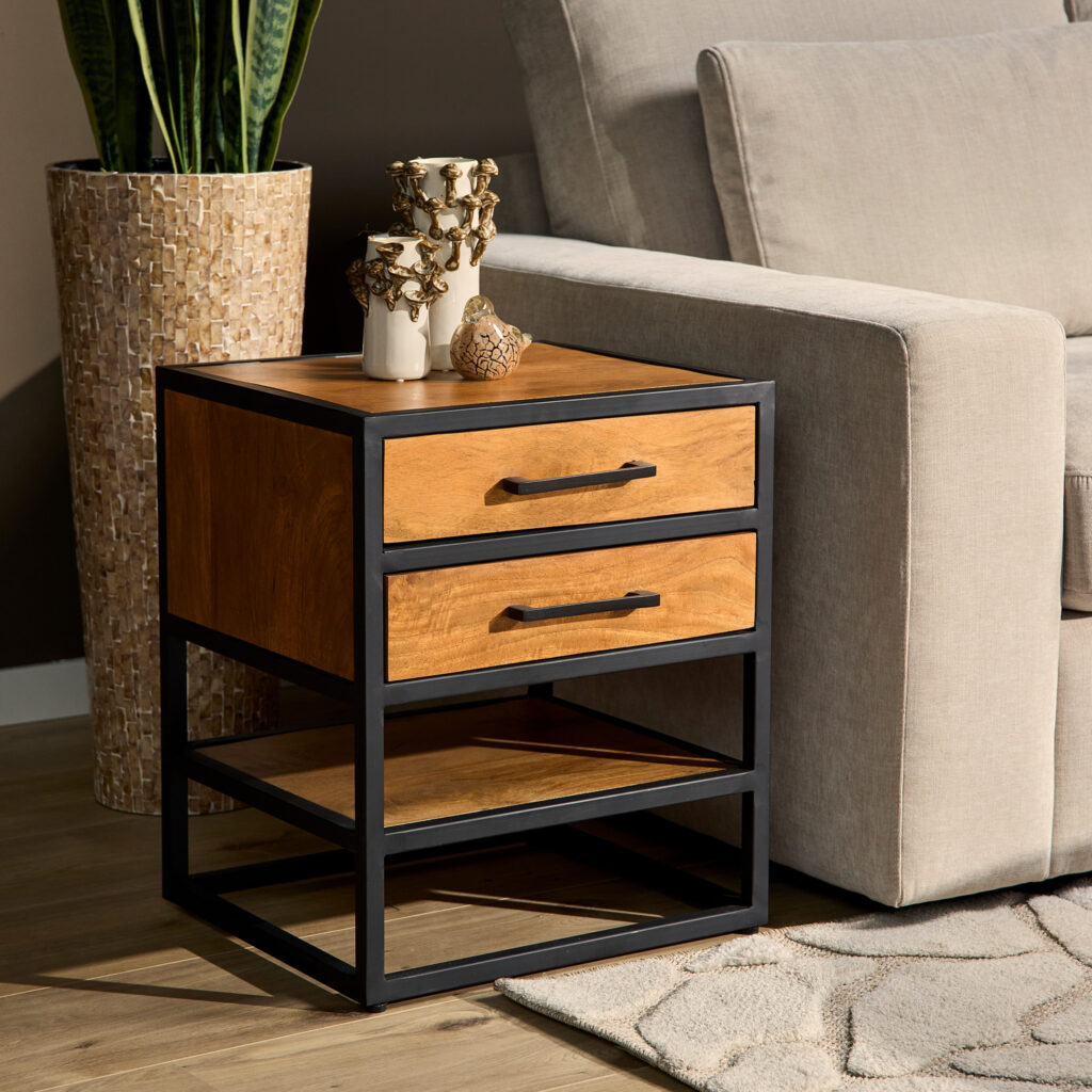 De Bijzettafel/nachtkastje Denver, gemaakt van mangohout en metaal met twee lades en een open plank, staat naast een beige bank. Op het tafeltje staan decoraties en kaarsen, terwijl op de achtergrond een hoge plant in een mozaïekpot te zien is.