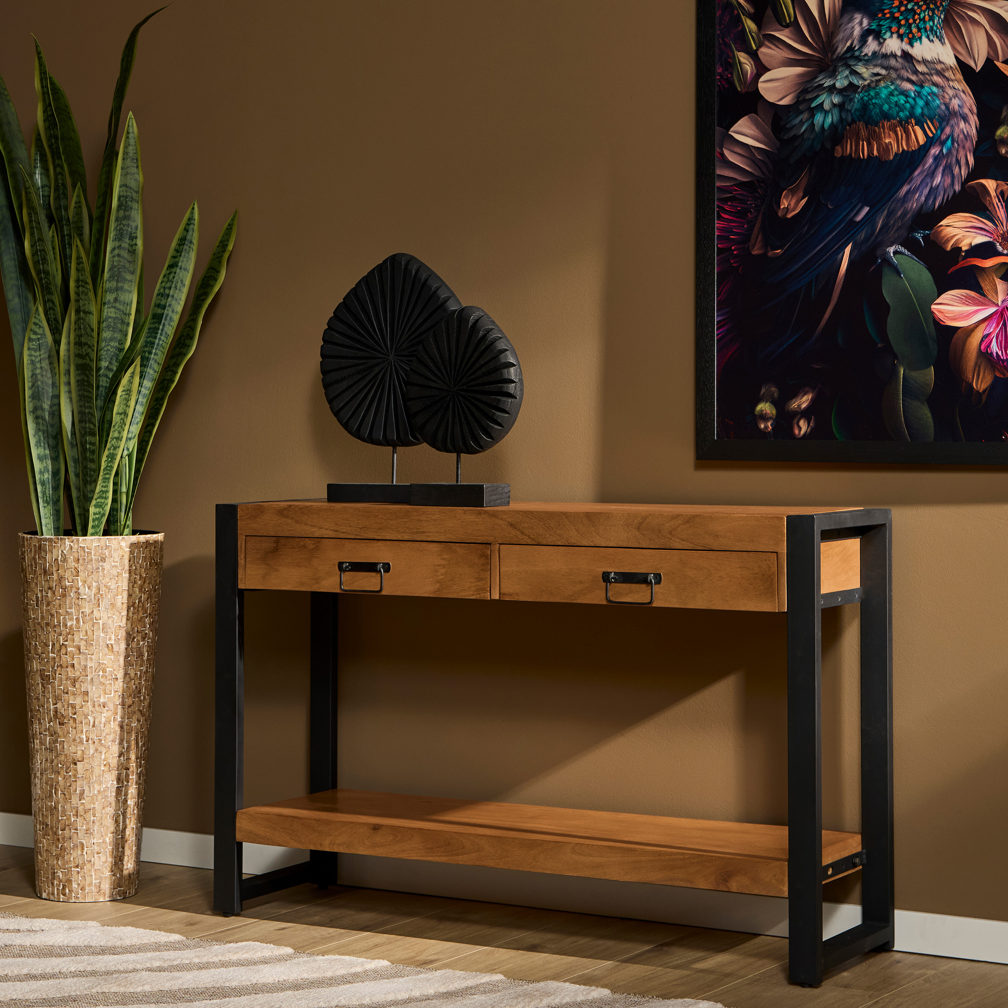 De Sidetable Boaz | 120 cm heeft een houten blad, een zwart metalen frame, twee laden en een onderste plank. Geplaatst tegen een bruine muur in de buurt van een hoge potplant en een vogelkunstwerk, is hij vormgegeven met een zwart decoratief beeld bovenop.