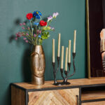Op een houten dressoir staat de Vaas Lyra - Antiek goud | Large met levendige kunstbloemen en een zwarte vijfarmige kandelaar met gouden kaarsen, tegen een groene muur naast ingelijste kunstwerken.