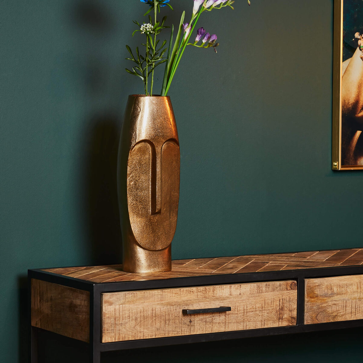 De Vaas Liv - Antiek goud | Large, gevuld met bloemen, staat op een houten consoletafel met zwart metalen poten en laden, tegen een donkergroene muur met rechts gedeeltelijk zichtbaar ingelijst kunstwerk.