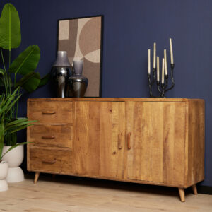 De Dressoir Omaha | 180 cm staat tegen een donkerblauwe muur. Bovenop staan twee vazen, een ingelijst abstract kunstwerk, een kandelaar met kaarsen, terwijl er een grote bladplant vlakbij staat. Het heeft drie laden en twee deuren.