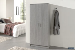 Een moderne slaapkamer met een keurig opgemaakt bed en de Opbergkast 'Ray' 2 deuren en 4 pootplanken Grijze eik (1c), voorzien van grijze deuren en handgrepen. Een grijze badjas hangt in de buurt en natuurlijk licht stroomt door een groot raam de neutraal getinte kamer binnen.