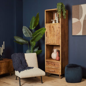 Een gezellige kamer met een beige stoel met een donkere deken, een hoge groene plant, de Boekenkast Omaha 55 cm houten kast met decoratiespullen, een blauw voetenbankje en moderne abstracte kunst op een marineblauwe muur.
