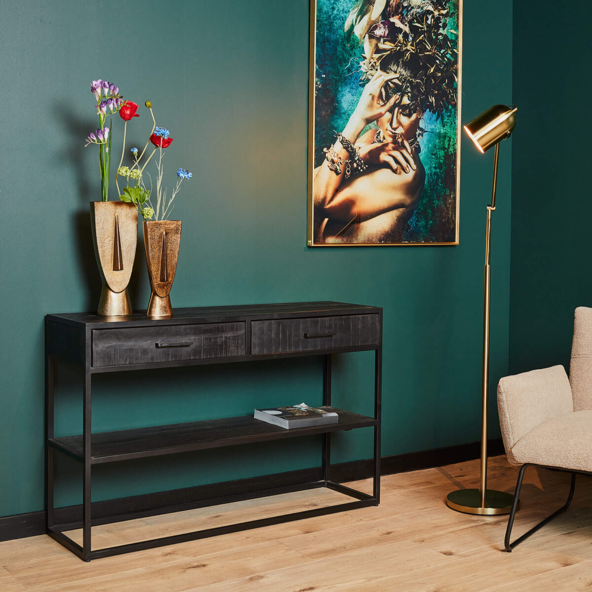 Een moderne woonkamer met de Sidetable Denver Zwart met 2 laden, gouden vazen met bloemen, een gouden staande lamp, een abstract portret op een groene muur en een beige stoel op een lichte houten vloer.