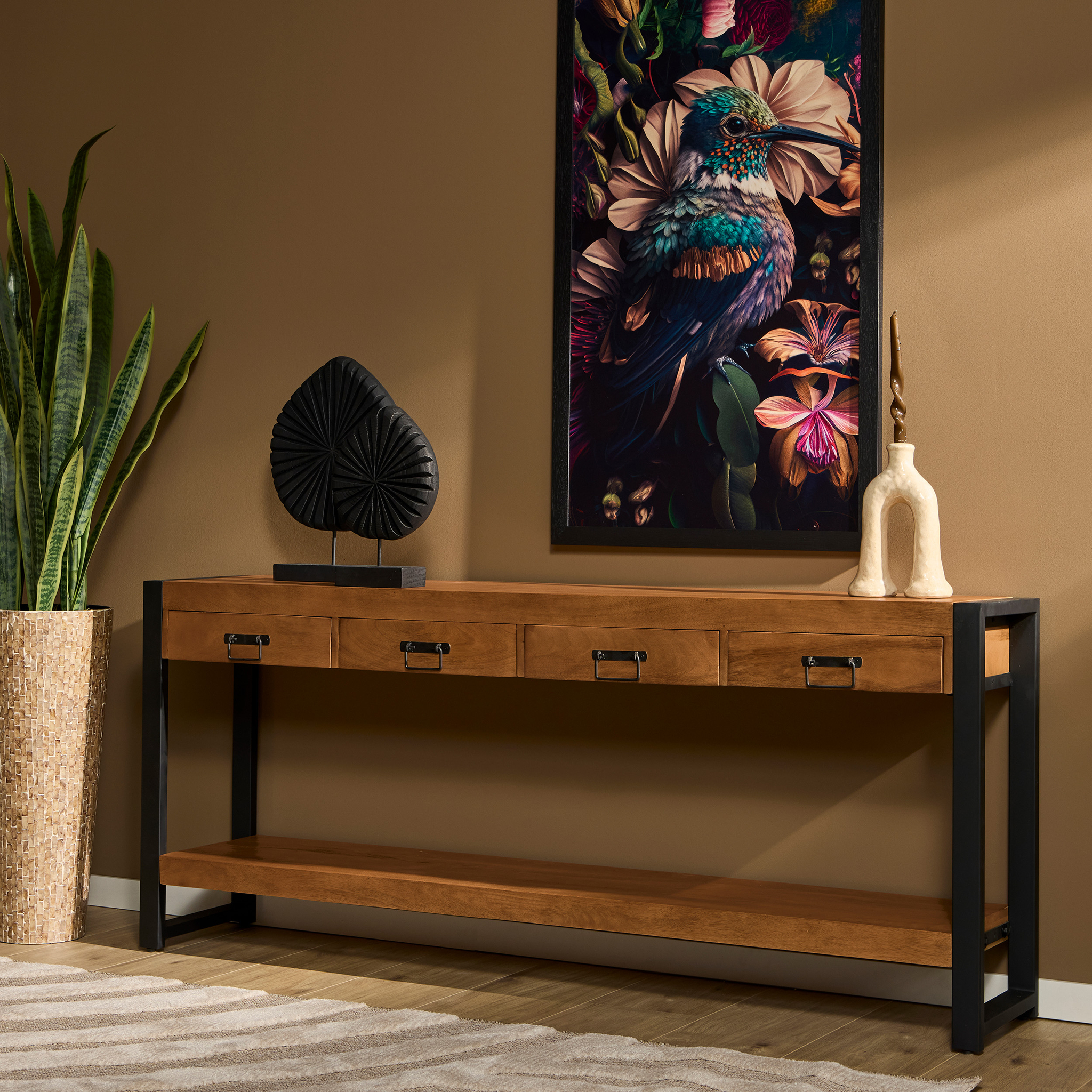 De Sidetable Boaz | 180 cm met zwart metalen poten staat tegen een gebruinde muur, versierd met abstracte sculpturen. Erboven hangen levendige vogelkunstwerken, terwijl links van de console een hoge potplant staat.