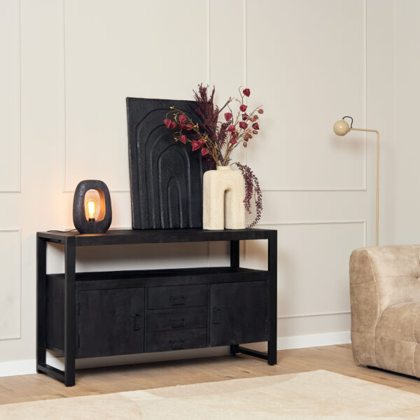 De Dressoir Boaz Black | 135 cm is vormgegeven met een brandende lamp, abstract zwart kunstwerk, een crèmekleurige vaas met droogbloemen en een neutrale inrichting in een lichte kamer met beige muren en een fauteuil.