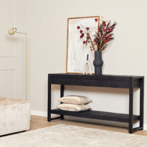De Sidetable Boaz Zwart | 150 cm staat tegen een beige muur, gestyled met een vaas gedroogde bloemen, een ingelijste foto en glasdecor. Op de onderste plank liggen twee decoratieve kussens. Vlakbij staan een beige bank en een staande lamp op een licht vloerkleed.